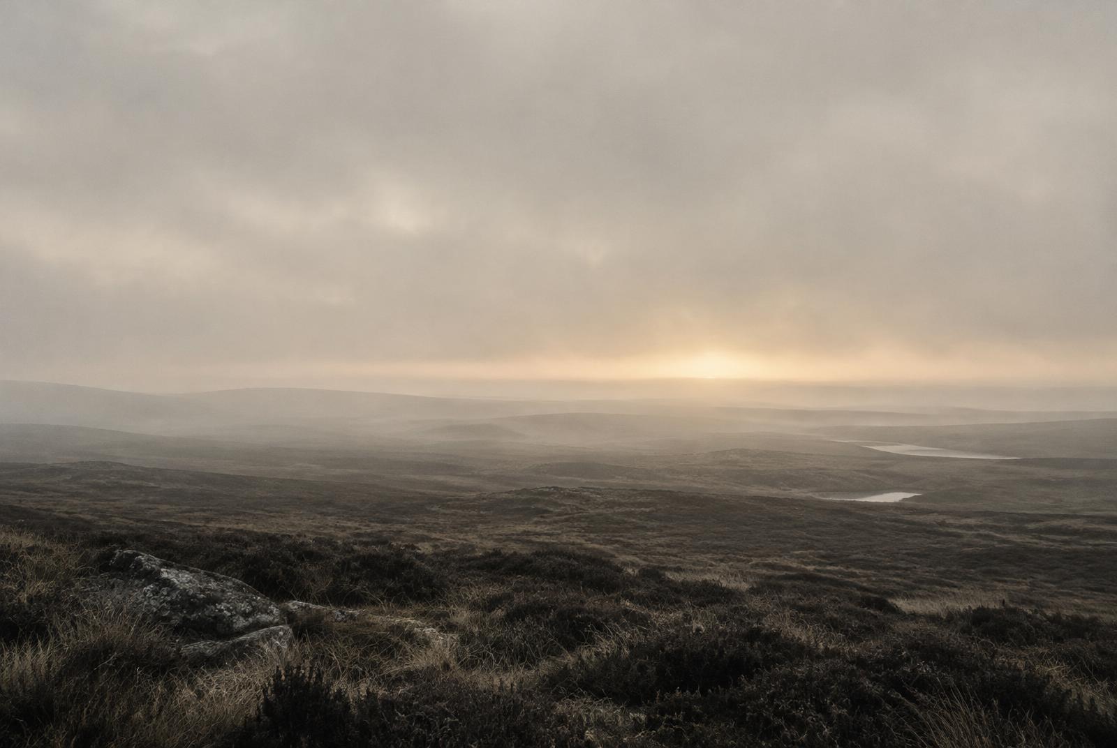 A vast empty landscape under fog, with a faint warm glow at the horizon